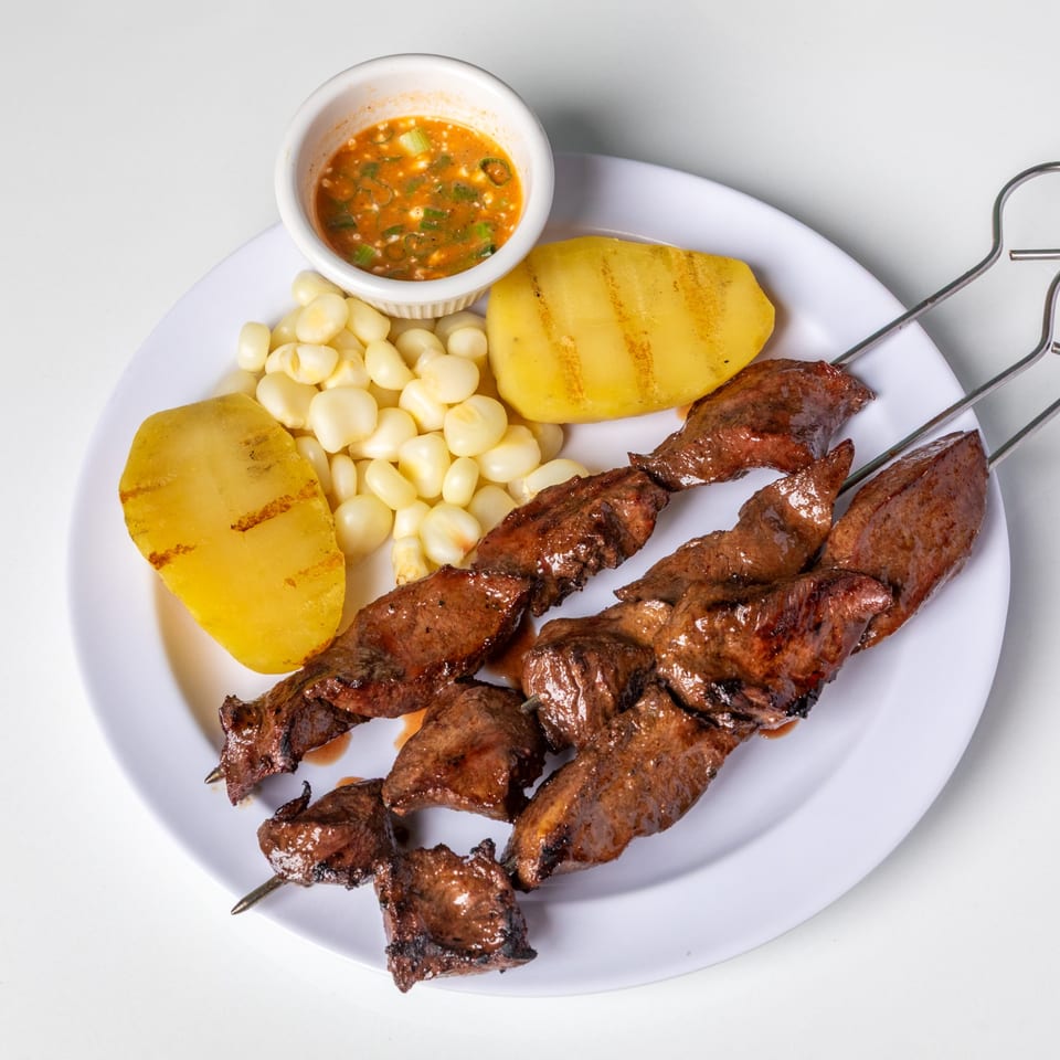 Two skewers of grilled anticuchos, with boiled corn, grilled potatoes, and a small bowl of sauce.