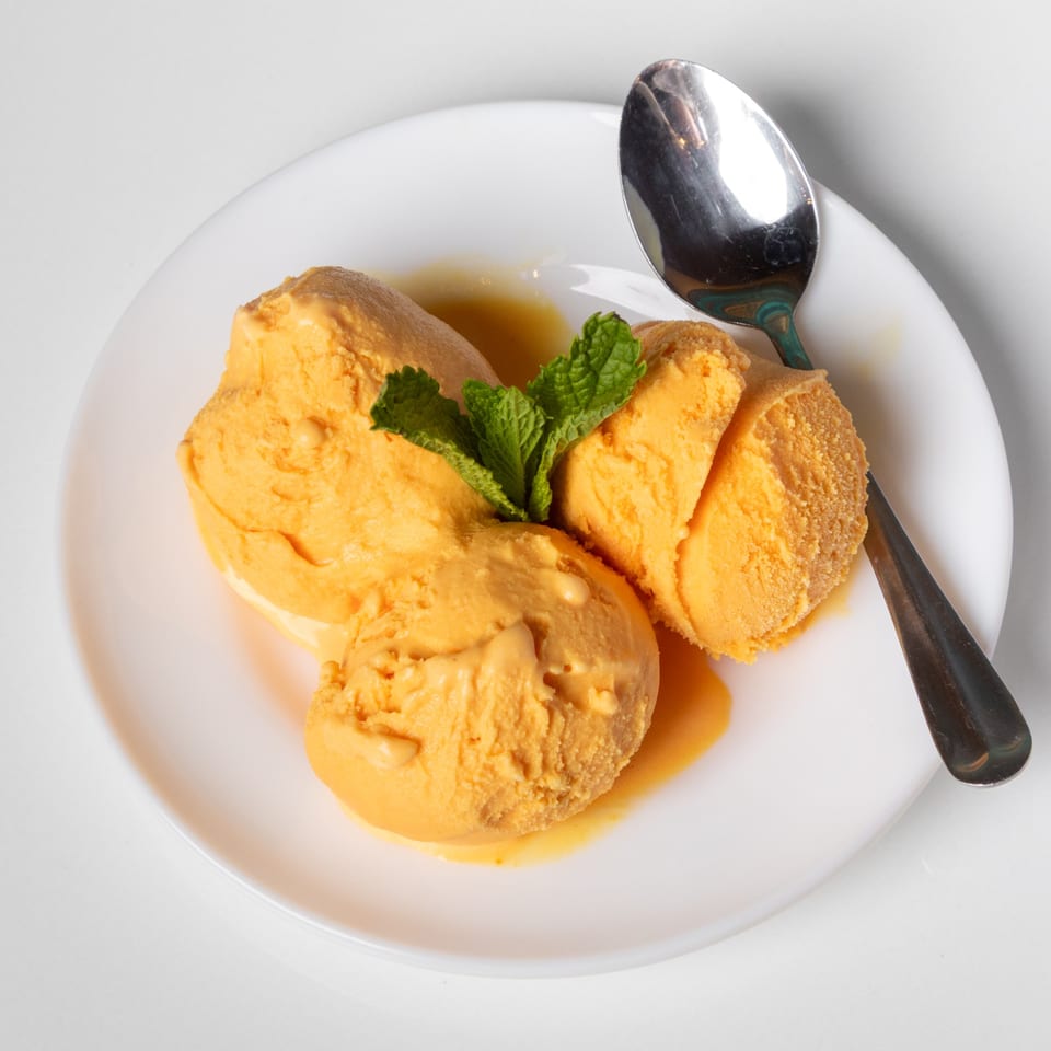 Three scoops of orange ice cream with mint garnish and a spoon on a white plate.